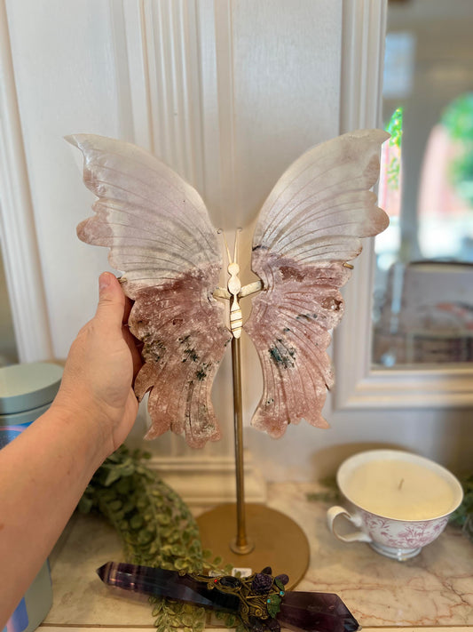 Butterfly-shaped crystal wings on a table with a hand showing the sizing of it, near a teacup and decorative items.